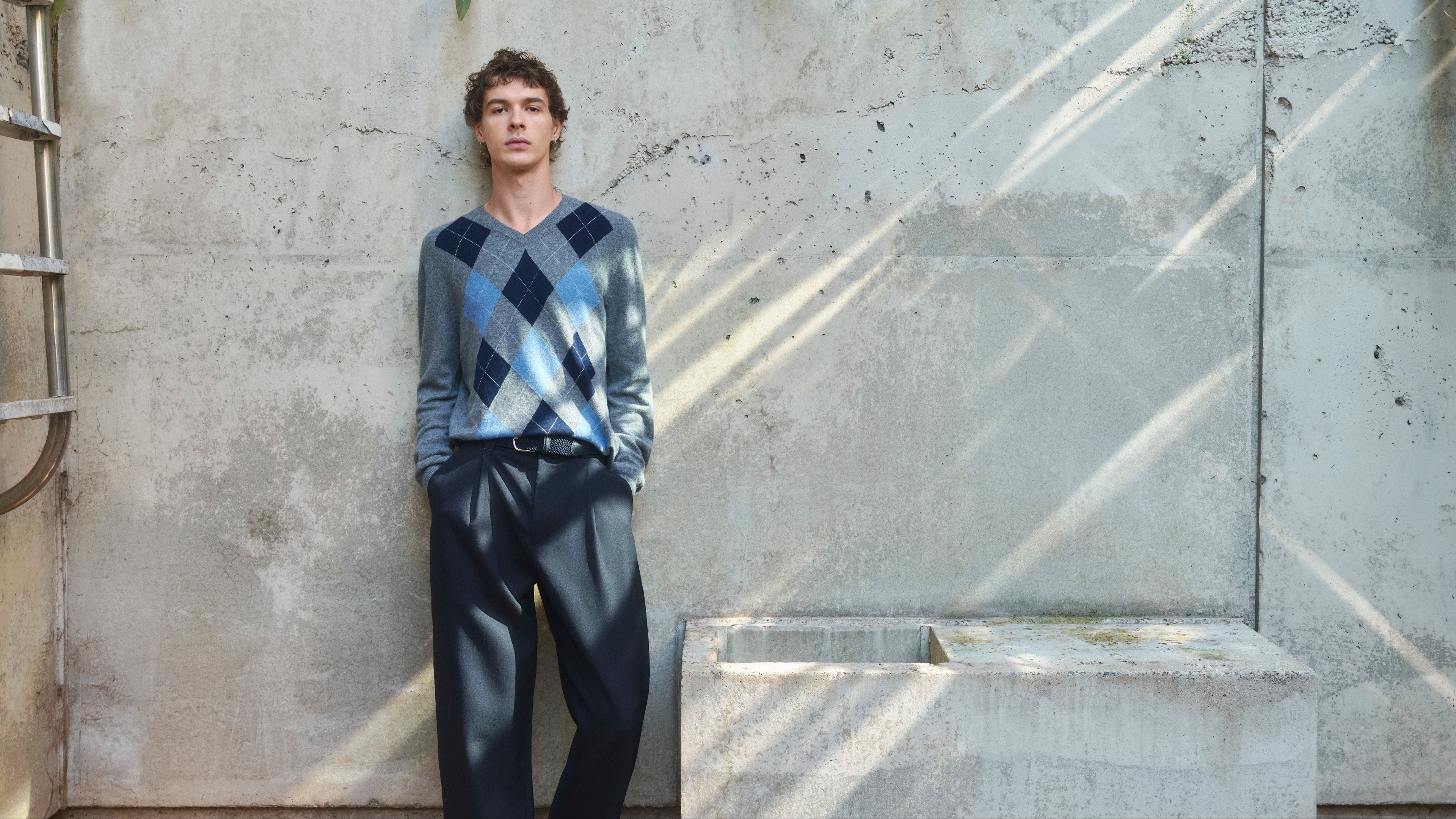 Man leaning against a concrete wall wearing a Pringle of Scotland Argyle cashmere sweater