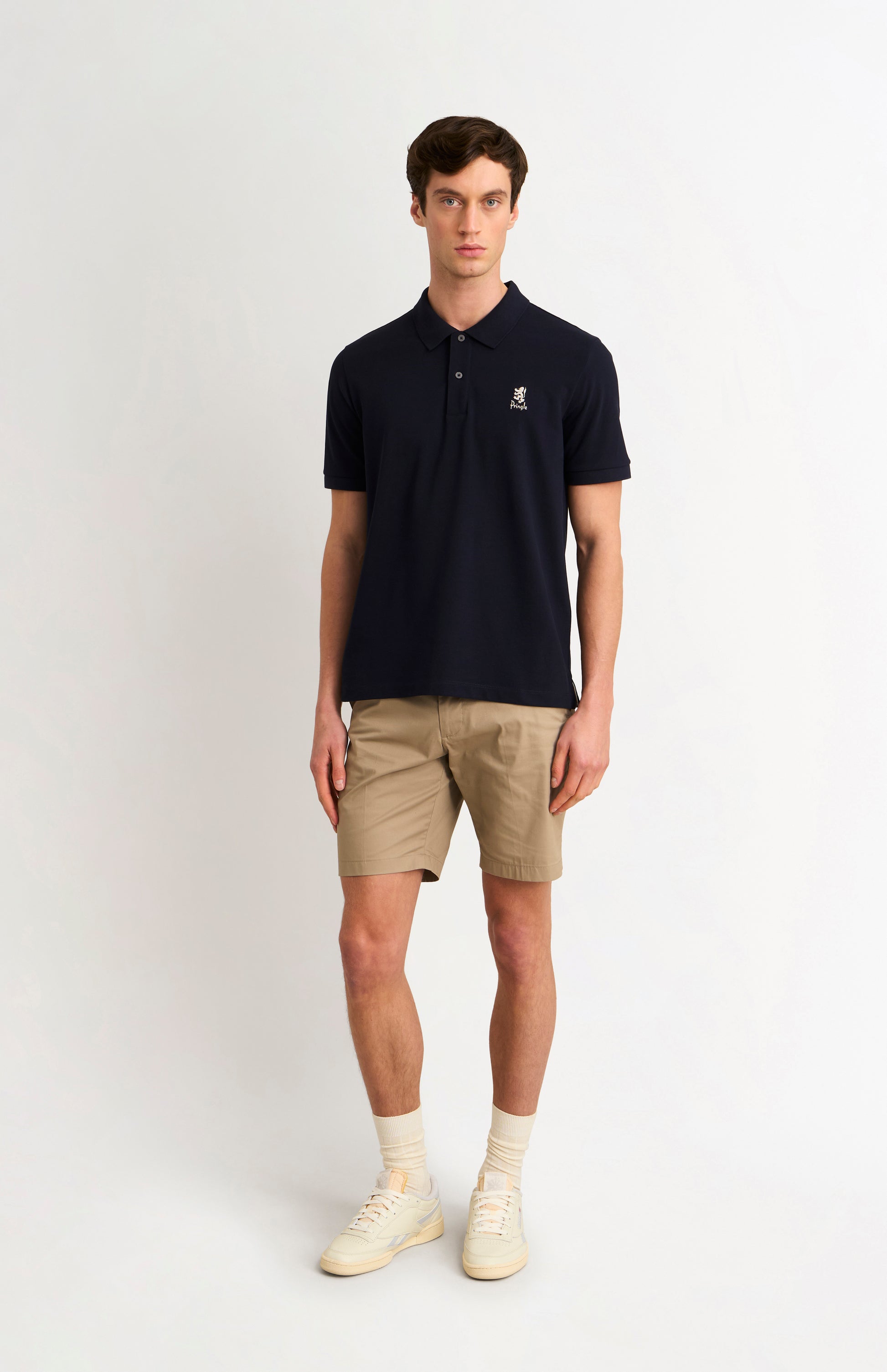 Man wearing a navy polo shirt and beige shorts on a white background