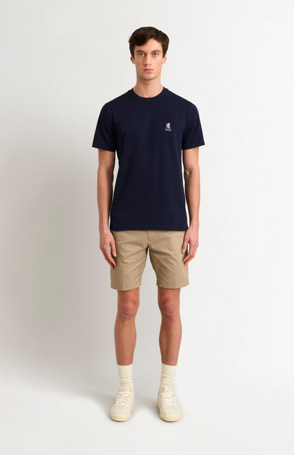 Man wearing a navy t-shirt with a small logo and beige shorts on a white background
